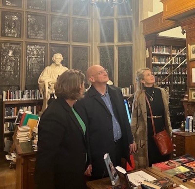 A man looks up in a college library. There is a statue behind him, and two women stand, one on each side of him, also looking up.