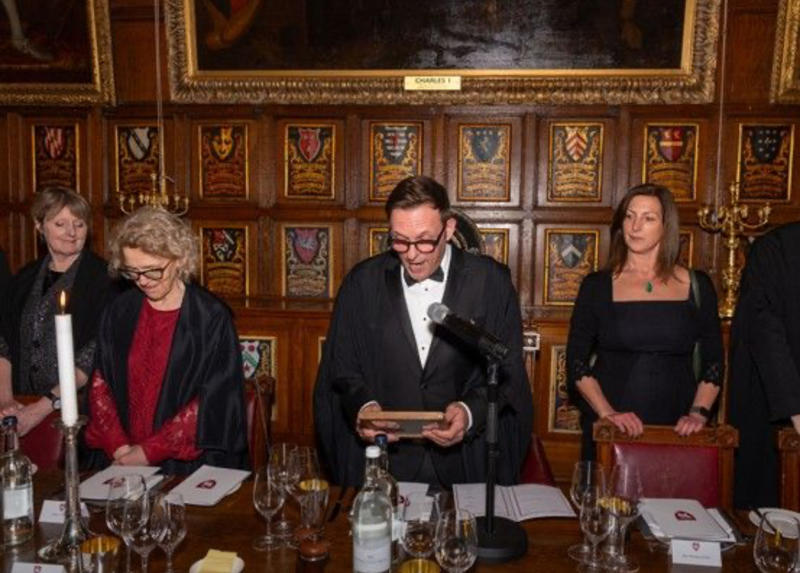 A man in glasses reading in a wood-panelled room with crests and portraits hanging on the wall, with three women on either side