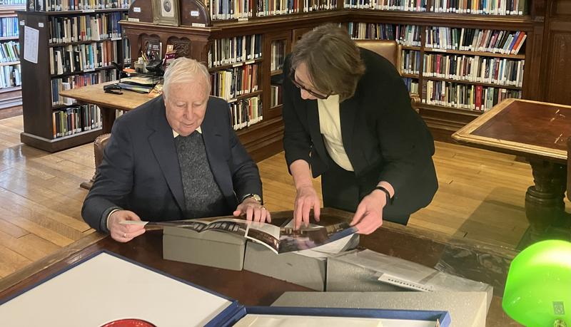 An elderly man sitting down inspecting materials in a library with a woman standing over him