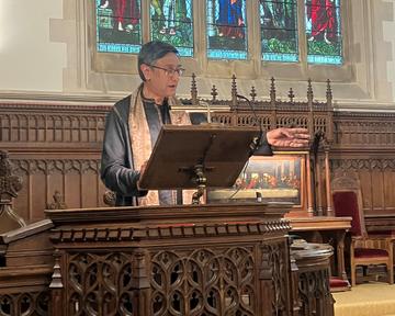 Prof Santanu Das speaking at the lectern of the college chapel with one hand outstretched