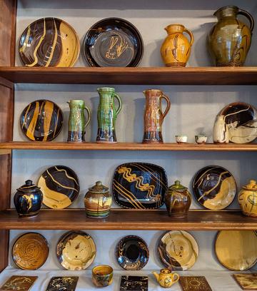 Four shelves of studio pottery on display with a range of shapes from tiles to jugs to large plates