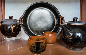 Two teapots and a plate in a dark glaze with two cups in the foreground