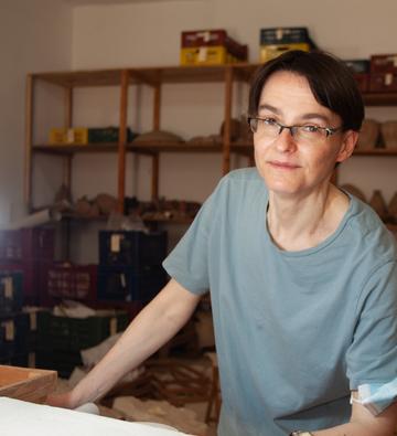 Photo portrait of Hana Navratilova, waring a t-shirt, with archaeological finds on shelves behind