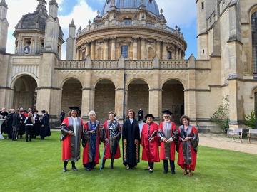 A line of women in scarlet doctoral robes, in the quad at All Souls College. They are standing on grass with a stone arcade and the dome of the Radcliffe Camera behind them.