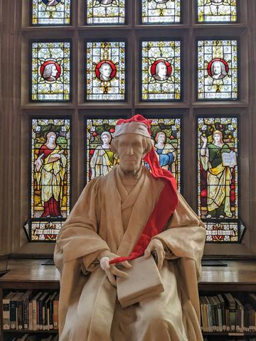 A picture of a marble statue of a man seated in academic robes.  The statue has a long Christmas hat placed on the man's head.