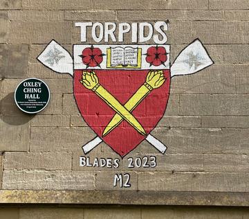 A painting on a wall next to a plaque with the words 'Oxley Ching Hall', showing a College crest with oars in the background and the words: 'Torpids, Blades 2023, M2'