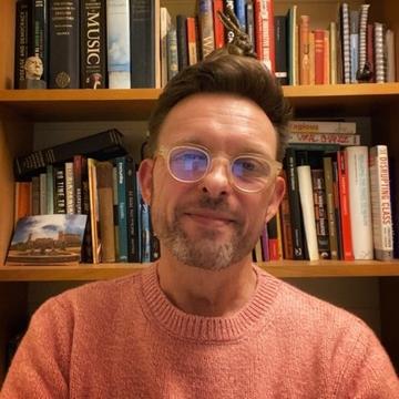 Portrait of Professor Matthew Weait smiling and facing directly ahead. He is wearing a light orange jumper and glasses, and is sitting in front of a bookcase.
