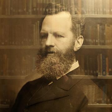 A photograph of a bearded man with shelves of books reflected faintly in the glass