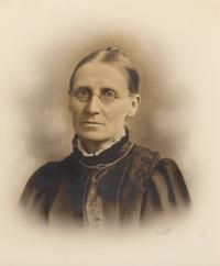 Black and white headshot photo of Lucy Toulmin Smith.  The sitter has her hair back, is wearing glasses and dressed in dark clothes.