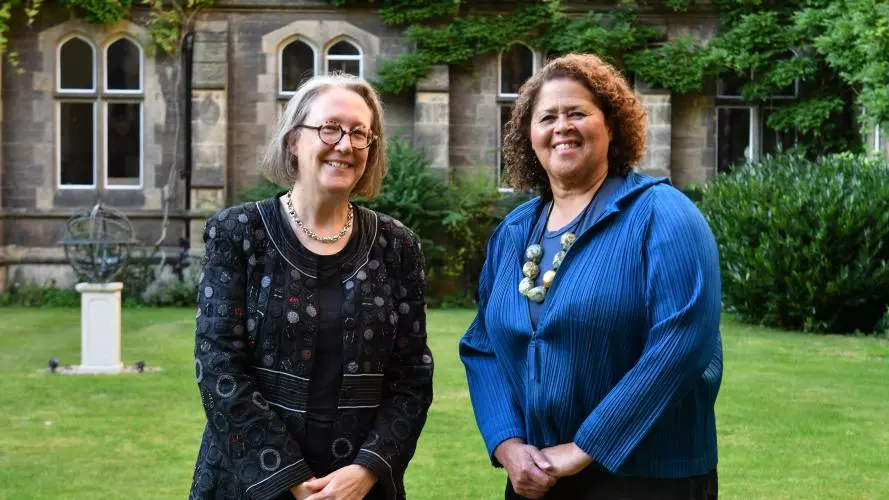 The Principal and Anna Deavere Smith smiling together in the Tate Quad. In the background, stonework, grass and green foliage
