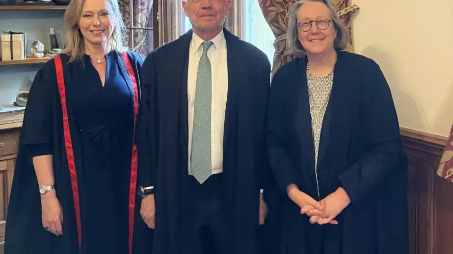 Three people, two women, one man in the centre, pose in academic gowns in a wood-panelled room