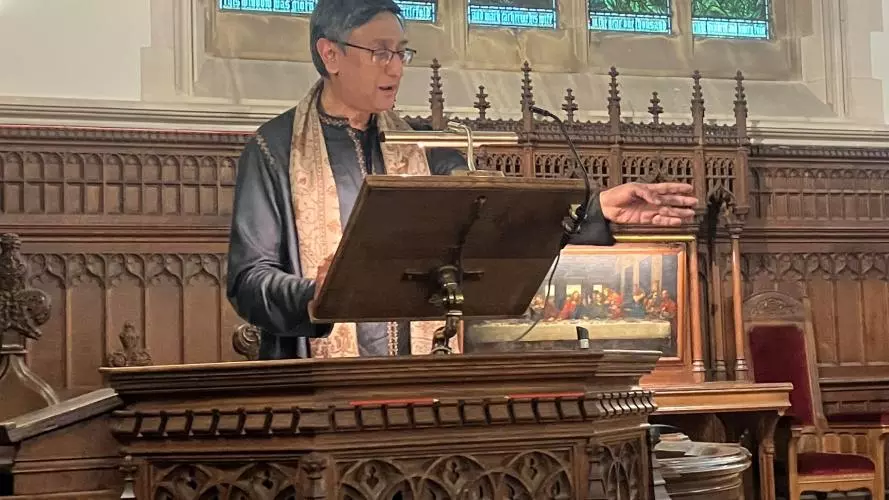 Prof Santanu Das speaking at the lectern of the college chapel with one hand outstretched