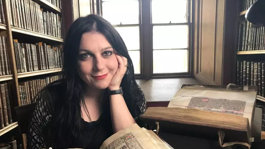Janina Ramirez posing in front of an old book with bookshelves and windows behind her back.