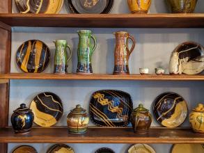 Four shelves of studio pottery on display with a range of shapes from tiles to jugs to large plates