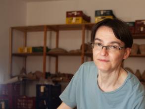 Photo portrait of Hana Navratilova, waring a t-shirt, with archaeological finds on shelves behind