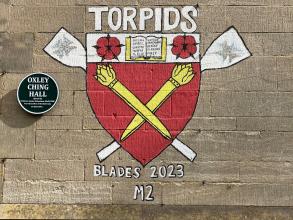 A painting on a wall next to a plaque with the words 'Oxley Ching Hall', showing a College crest with oars in the background and the words: 'Torpids, Blades 2023, M2'