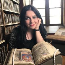 Janina Ramirez posing in front of an old book with bookshelves and windows behind her back.