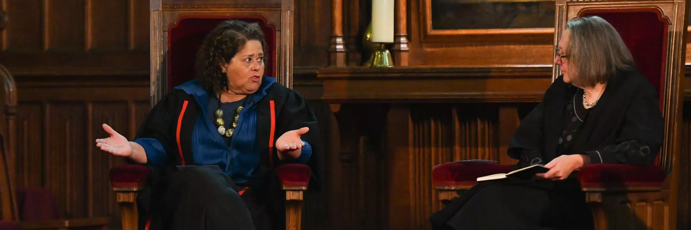 The Principal and Anna Deavere Smith are seated together in high-backed chairs at the front of the Chapel. Anna is on the left, gesturing as she explains a point, while the Principal (on the right) listens attentively. 