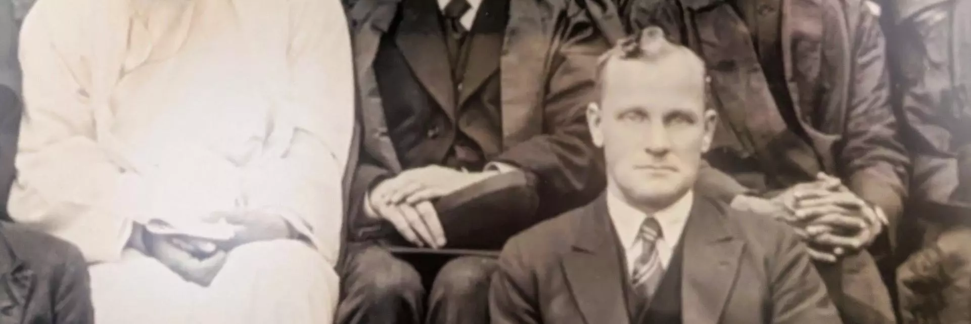 A detail of a photograph showing four figures. Three are seated next to each other on chairs and show from left to right, a man with a white beard and white robes, and two men in suits and academic gowns, one wearing a turban. 